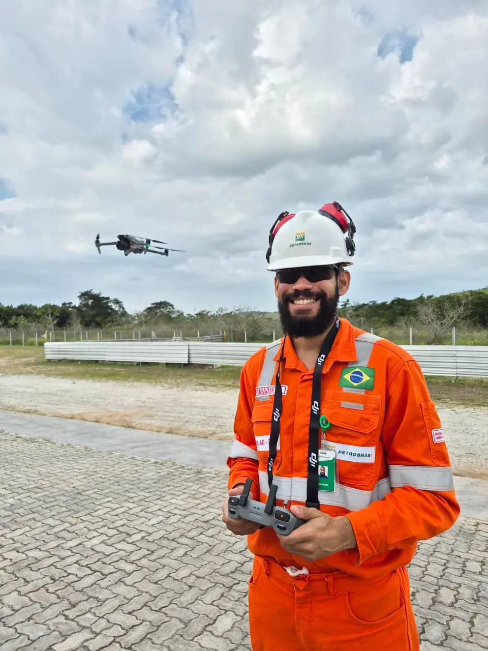 Aluno de macacão laranja pilota o drone em área externa durante o treinamento, com a aeronave no ar ao fundo