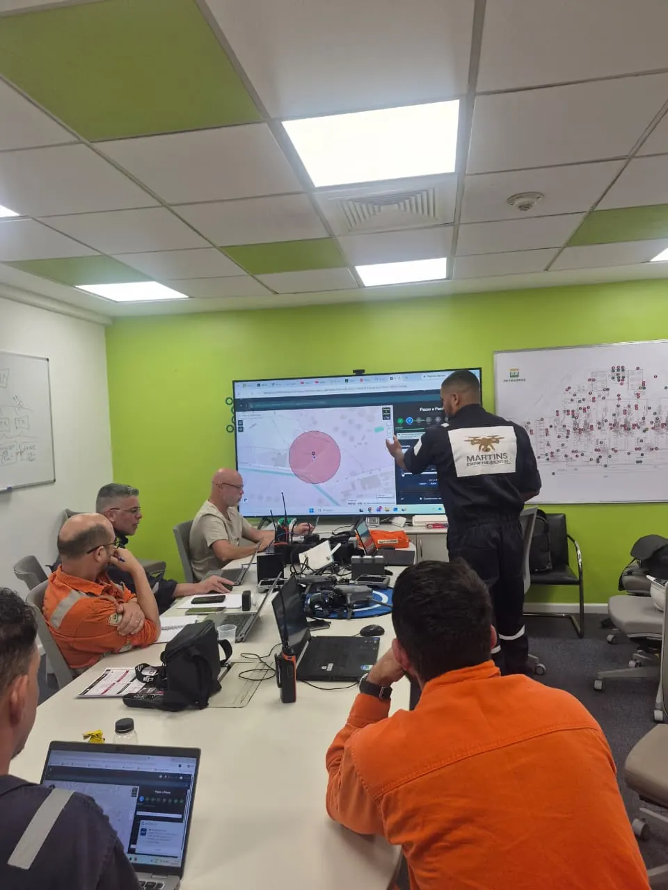 Turma em sala de aula acompanha o instrutor, que aponta para um mapa com uma área de voo demarcada na tela