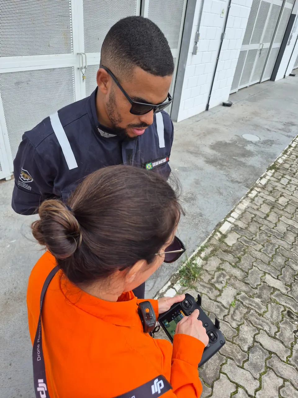 Instrutor acompanha de perto uma aluna que opera o controle do drone em área externa