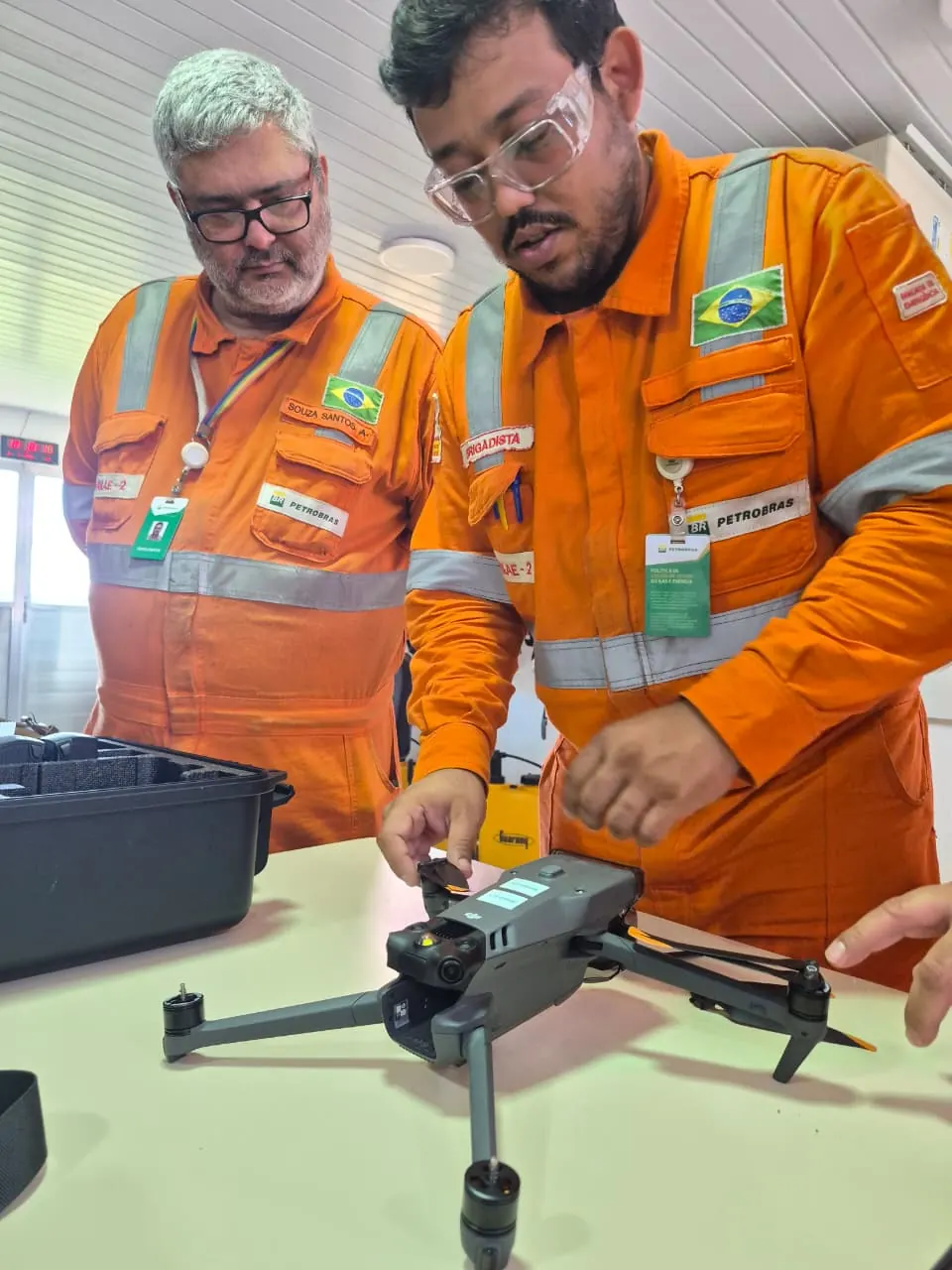 Dois alunos de macacão laranja preparam um drone sobre a mesa, ao lado da maleta do equipamento