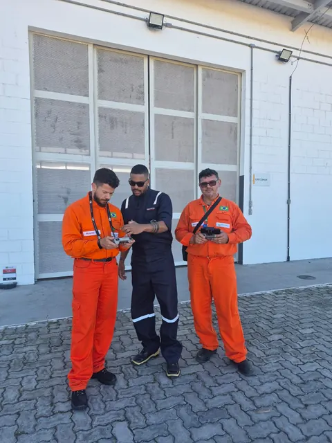 Instrutor entre dois alunos de macacão laranja, todos com os controles do drone em mãos